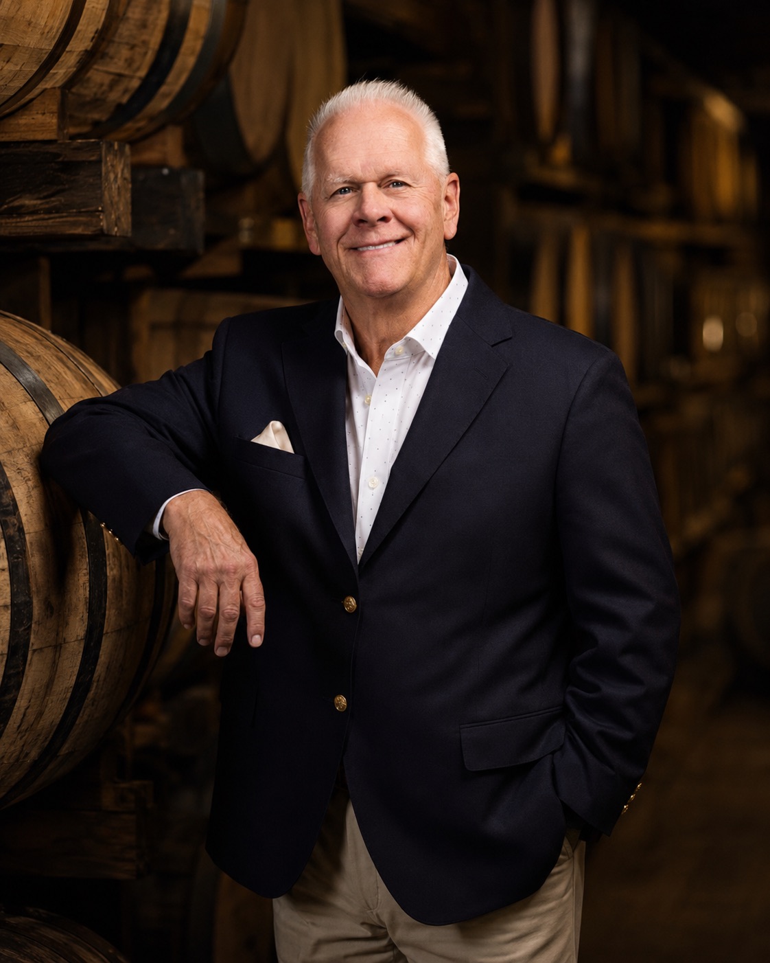 Chip Flanders in a navy blazer, leaning on a whiskey barrel in a barrel-aging warehouse.